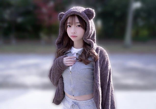 クマ耳フードの女性が公園のような屋外で立ち、グレーの服に手を添える